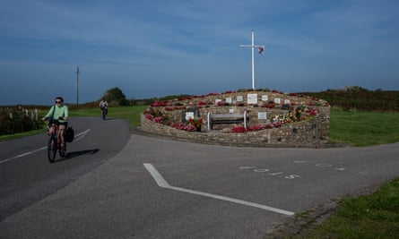 A woman with sunglasses and no bike helmet cycles along a fork in an Alderney road at a memorial with plaques, flowers and a tall thin cross