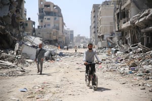 A man rides a bicycle near the debris of damaged building while another man walks along the dusty road