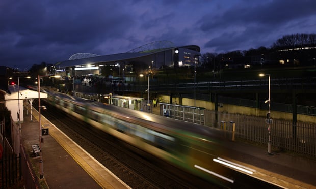 A train speeds through a station with a football ground in the background.