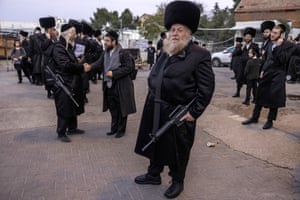 A bearded man wearing a black fur hat stands in the foreground carrying an M16, as another armed man shakes hands with a person in the background. Other Jewish men wearing traditional outfits stand around them