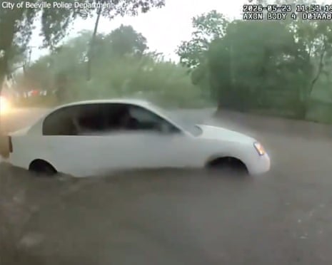 A white sedan is submerged up to its wheel arches in floodwater on a street
