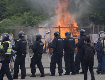 Police in riot gear stand in a line while a large fire burns in the background.