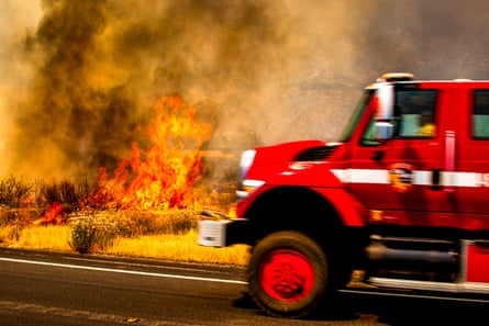 An emergency vehicle driving along a road as flames and smoke rage in the background. An emergency vehicle driving along a road as flames and smoke rage in the background.