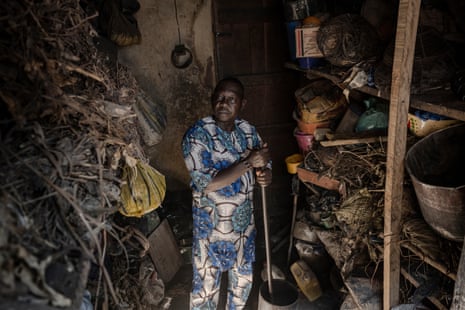 A Nigerian man wearing matching print dress shirt and trousers stands in a room lined with shelves stuffed with dried herbs and pots. He holds a long pole to mix something in a large bowl