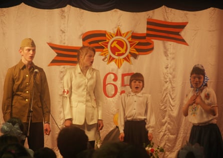Four children in costumes stand on a stage with a white backdrop with a hammer and sickle symbol and ‘65’ stitched on