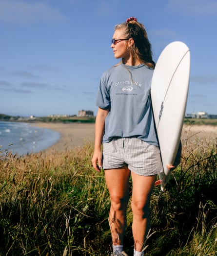 She stands with a surfboard on the headland above a beach, wearing Quiksilver shirt and check shorts