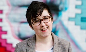 Head and shoulders photo of Lyra McKee outside the Sunflower Bar on Union Street in Belfast: she has short dark hair, wears glasses and red lipstick, a brown tweed jacket and striped top, and is smiling; a red, pink, blue and white graphic mural is seen behind her.