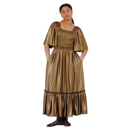 A model wears a long dark gold smock dress with elbow length sleeves and a square neckline. They also wear a chunky gold necklace and black patterened tights.