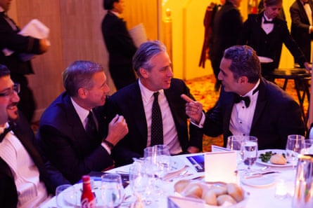 Three men in suits sit at a table talking to each other