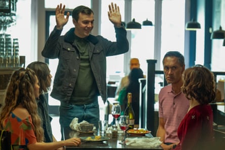 A man stands with his hands raised around a dining table, where four people are seated