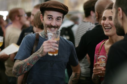 Two young men and a woman smile and hold glasses of beer at the festival in 2015; the man facing the camera has a hipster-style moustache, brown cap and heavily tattooed arms.
