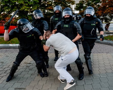 A group of police wearing armour and helmets, one of them swinging a baton, detain a man who is crouching in a defensive pose