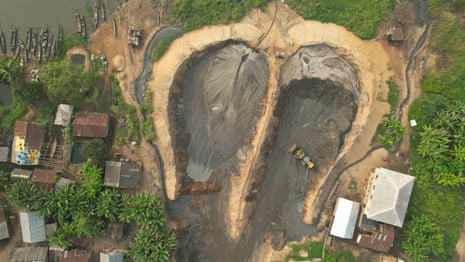 ‘The water is no longer our friend’: how dredging is pushing Lagos Lagoon towards ecosystem collapse – photo essay Drone view of a dredging site shows two deep channels dug out of the ground next to a river. Heavy machinery spews dark slurry near residential houses and dense greenery.