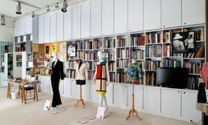 A display at the Musée Yves Saint Laurent in Paris. Mannequins wear a black trouser suit, a 1960s geometric-pattern colourful dress and also a white jacket, and stand in front of a wall of books and magazines.