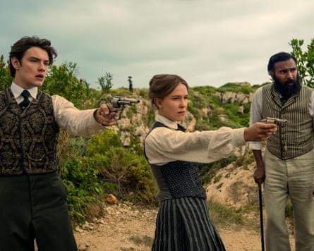 Louis Partridge as Tewkesbury, Millie Bobby Brown as Enola Holmes, pointing a gun, while Himesh Patel as Dr Watson looks on in Enola Holmes 3.