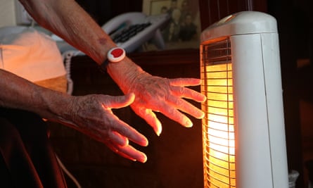 An elderly woman warms her hands at an electric heater.