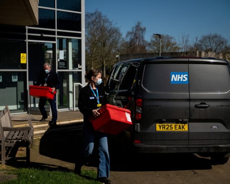 Two masked NHS workers carry red boxes to a van outside a modern building