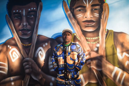 A man in a colourful outfit and baseball cap stands in front of a wall painted with murals of people with painted faces and bodies, holding what look like clubs.