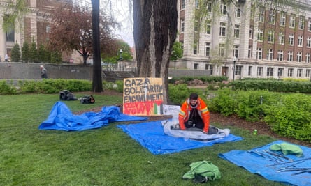 A man in an orange jacket kneels on a blue tarp and rolls up a tent, in front of a piece of plywood spray-painted with the words “Gaza Solidarity Encampment - divest now.”