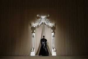 A model in a black outfit with their back to the camera walks under a large ornate silver arch and through curtains behind it