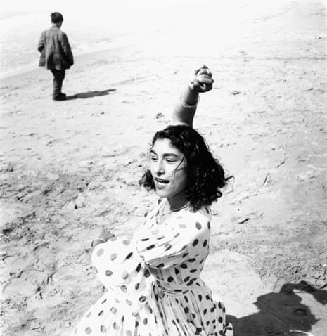 Woman in polka-dot dress dancing on a beach as a child walks behind her 