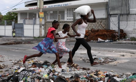 Children run across rubble holding hands with a woman who has a sack of rice on her head,