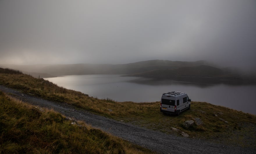 A solitary campervan parked overlooking a rugged lake landscape as sun breaks through heavy mist. 