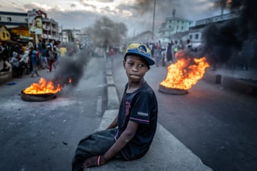 A boy sits atop a wall near two burning tyres