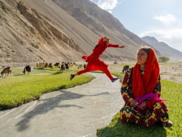 An Afghanistan woman sits in sunlit countryside whilst another jumps across a path behind her.