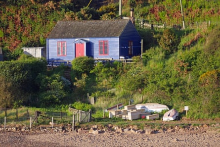 A blue timber cabin with red painted window frames by a rock shore.