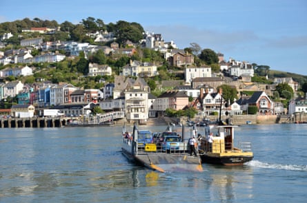 A small car ferry being pulled across a river by a tugboat, with houses going up a hill on the other side