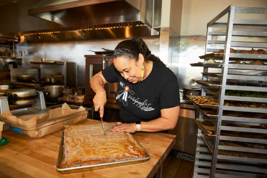 A woman cuts into a large pan of cornbread in an industrial kitchen. Baker’s racks on the right hold more pans of food.