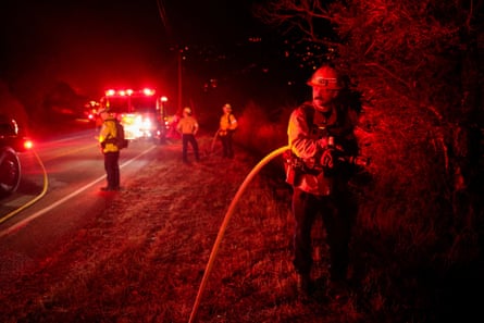 Firefighters stand beside road at night to work on fire, faces lit by the red lights of trucks