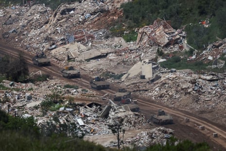 A line of six Israeli tanks ride past piles of debris from destroyed buildings.