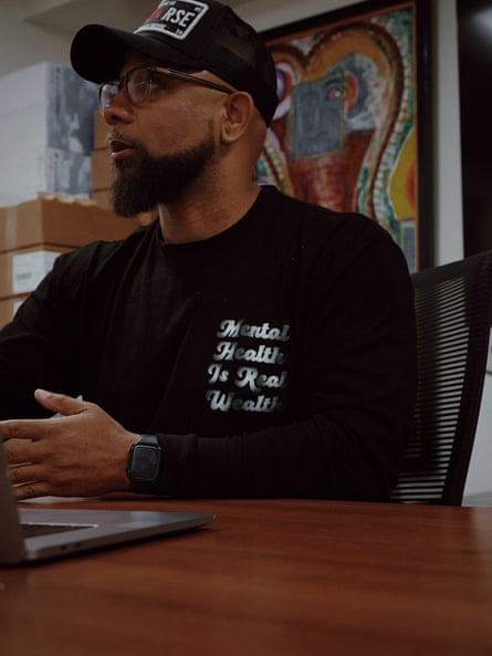 a man wearing a “mental health is real wealth” tee shirt speaks while seated at a table