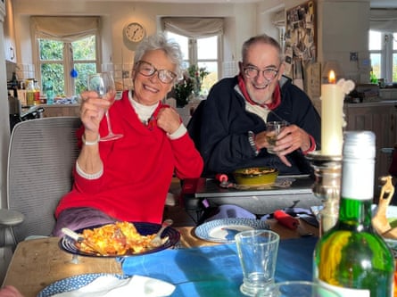 Sarah in a red jumper holding up a glass of wine and Michael in a dark top, holding a glass, sitting at a table together in a kitchen with big windows behind them.