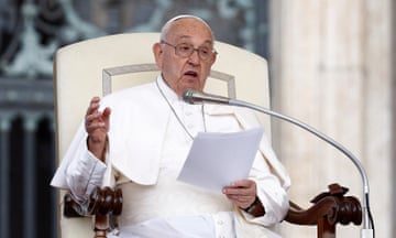Pope Francis seated in white holding papers, with microphone in front