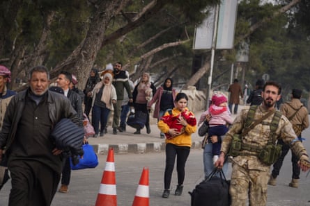 Men, women and children walk along a pavement and roads, including a man in military uniform carrying a bag