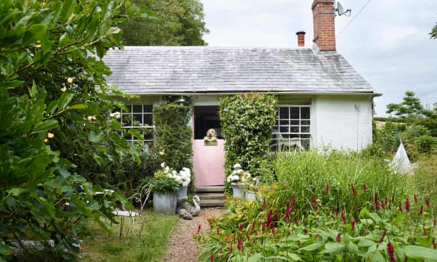 Single storey cottage with a pink stable door in the middle with a dog peering over the top, a path leading up to it and an abundant garden