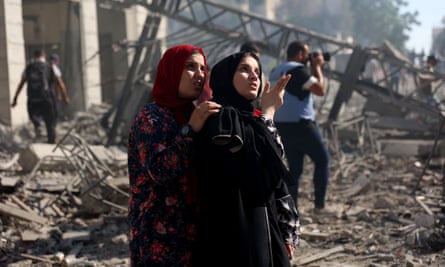 two young Palestinian women, one with her arms around the other’s shoulders, are looking upward at the damaged building; they look shocked and saddened. They both wear headscarves, one dark red over a patterned robe, the other’s black over a black robe. A man is seen behind them standing amid the rubble taking a photograph; debris and sections of destroyed structures are seen in the background. 