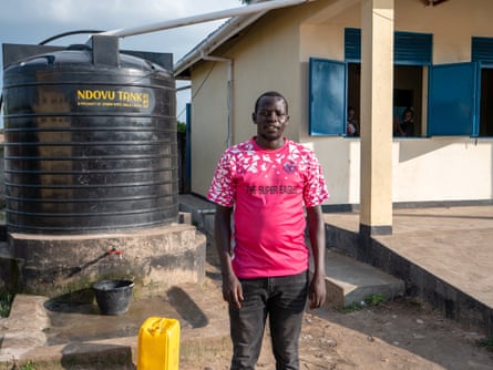 A man stands in front of a water tank and a building with blue shutters on its windows