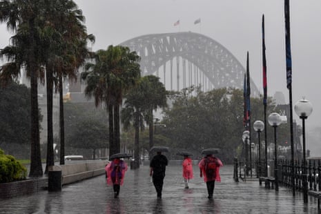 People wearing ponchos and holding umbrellas walking along the harbour in Sydney in rain with the harbour bridge in the background