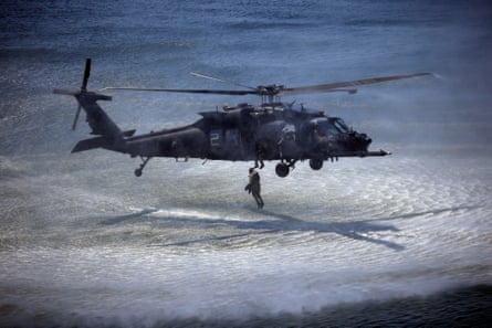An MH-60 Black Hawk hovers over the sea as a soldier jumps from a hatch; the helicopter and its rotors cast shadows over the water.