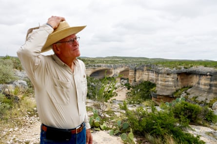 An older man in a cowboy hat stands near a canyon in the countryside.