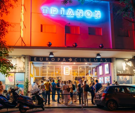 The night-time exterior of a cinema, with blue neon sign on an orange wall and people mingling outside the entrance.