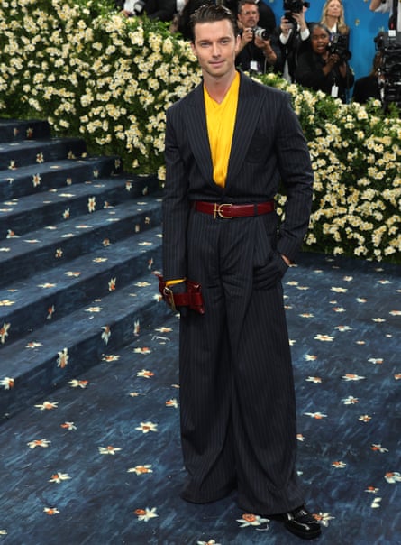 The celebrity Patrick Schwarzenegger in a two piece navy suit brown belt and yellow top. He holds a brown clutch and is on a blue carpet with a white daisy pattern. There are flowers and photographers in the background.