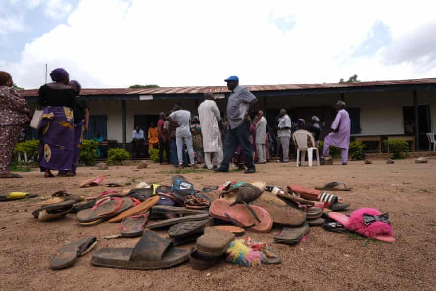 A pile of flipflops in the yard of a school with people milling about in the background