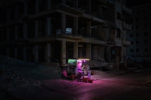 Men sit outside a kiosk in front of a damaged building in dark