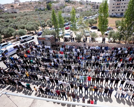 A high wide shot of hundreds of mourners standing in lines in an open square in the hilly village of al-Mughayyir