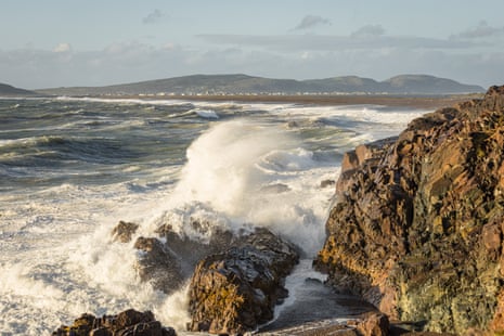 A wave crashing on a rocky shore with a long beach in the background and houses seen in the far distance